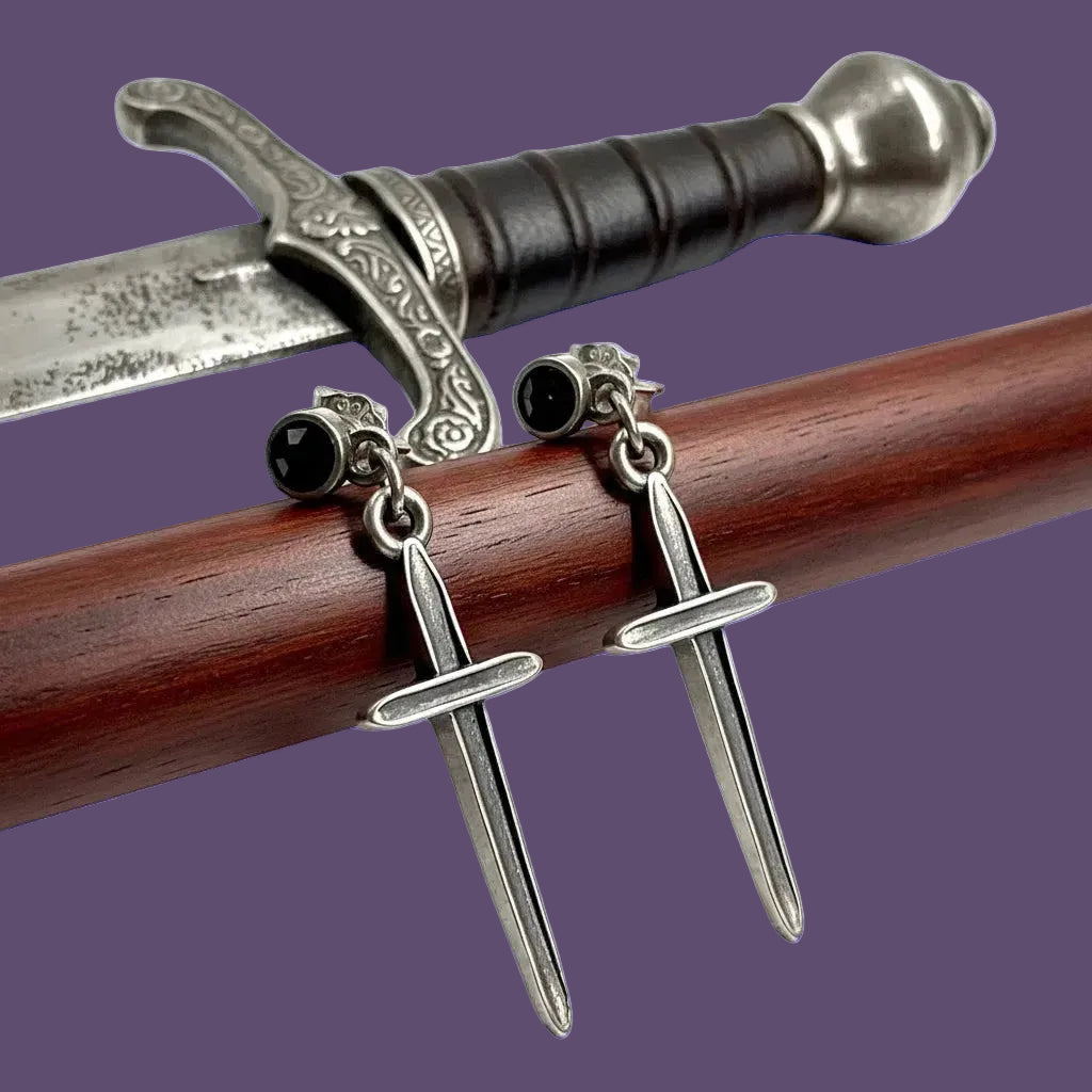 A pair of silver dagger-shaped earrings with black round studs, displayed on sword prop against a purple background.