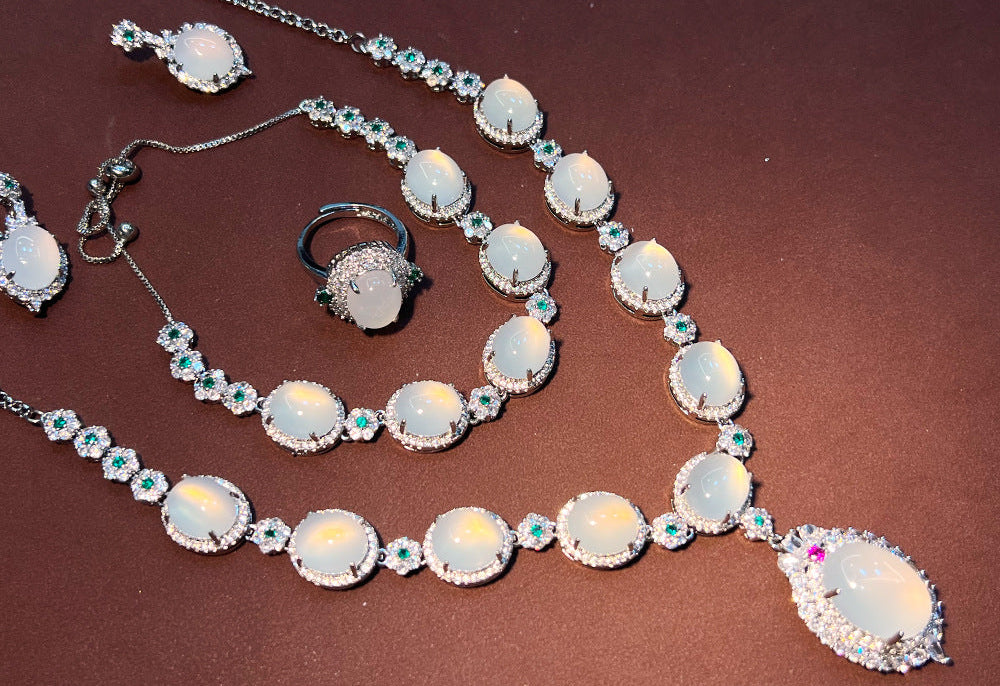 A silver-tone jewelry set featuring a necklace, bracelet, ring, and earrings with large oval milky-white stones and small turquoise-blue accent stones, arranged on a dark brown background.